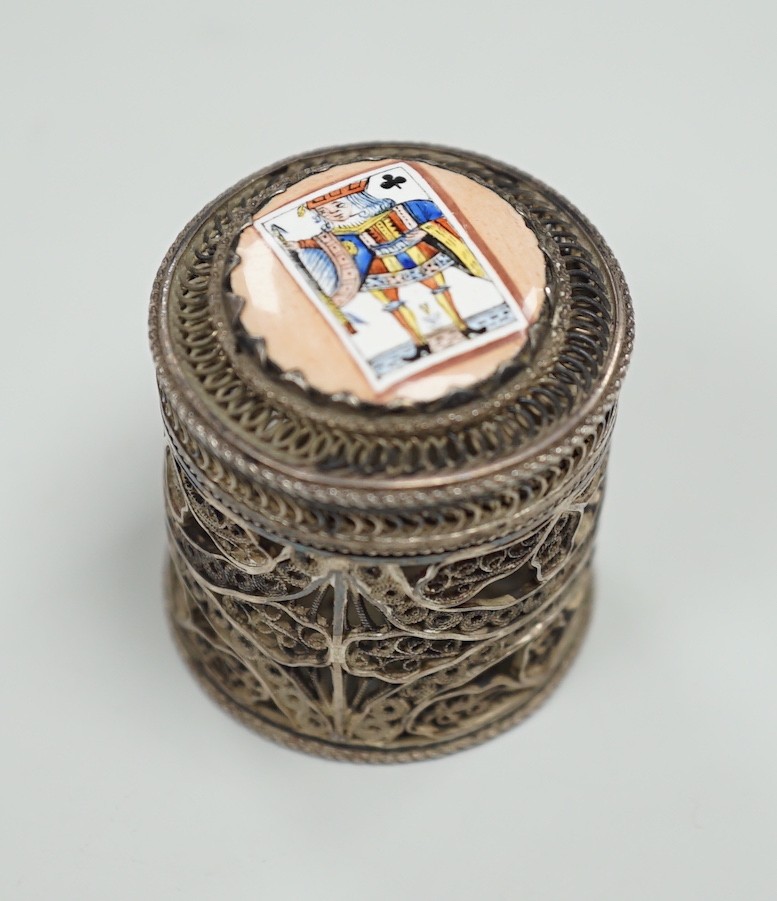 A continental filigree white metal and enamelled cylindrical pill box, the cover enamelled with a picture playing card, height 32mm.